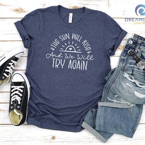 May include: A heather navy blue t-shirt with a white graphic that says "The sun will rise and we will try again". The graphic features a stylized sun with a heart in the center. The shirt is laid flat on a wooden surface with a pair of black sneakers and a pair of blue jeans.