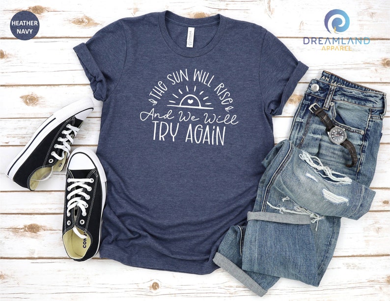 May include: A heather navy blue t-shirt with a white graphic that says "The sun will rise and we will try again". The graphic features a stylized sun with a heart in the center. The shirt is laid flat on a wooden surface with a pair of black sneakers and a pair of blue jeans.