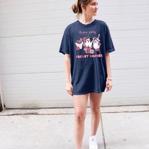 Pode incluir: Uma camiseta azul marinho com uma fita rosa e fantasmas com o texto "Scare away breast cancer".