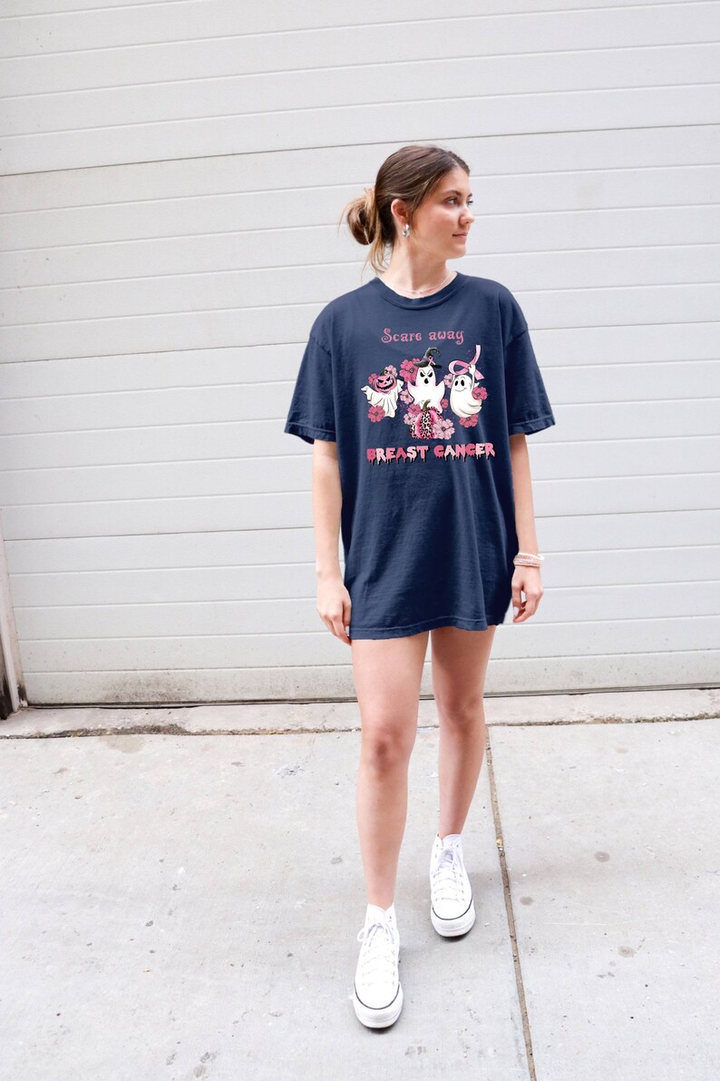 Pode incluir: Uma camiseta azul marinho com uma fita rosa e fantasmas com o texto "Scare away breast cancer".