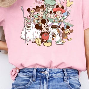 May include: Pink t-shirt featuring a colorful graphic of Disney characters, including Mickey Mouse, Woody, Buzz Lightyear, and others. The shirt has a crew neck and short sleeves. The text "Happy Place" is visible on the shirt.