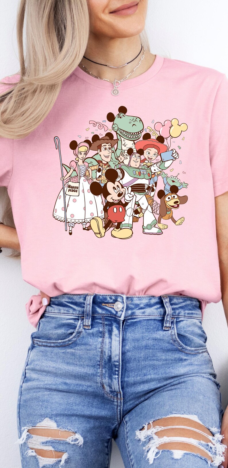 May include: Pink t-shirt featuring a colorful graphic of Disney characters, including Mickey Mouse, Woody, Buzz Lightyear, and others. The shirt has a crew neck and short sleeves. The text "Happy Place" is visible on the shirt.