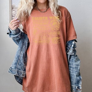 May include: A rust-colored oversized t-shirt with a gold graphic print that reads "Fourth Wing" and includes a quote about love and destiny. The shirt is worn by a person with blonde hair and a denim jacket.