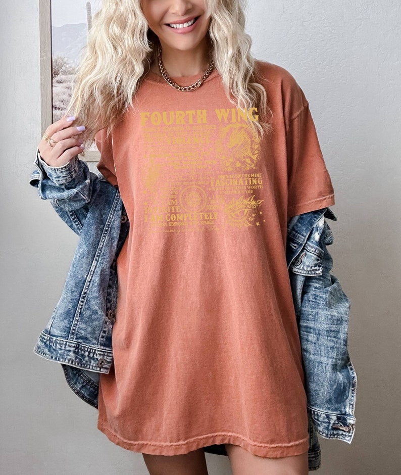 May include: A rust-colored oversized t-shirt with a gold graphic print that reads "Fourth Wing" and includes a quote about love and destiny. The shirt is worn by a person with blonde hair and a denim jacket.