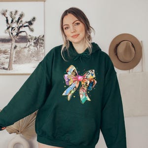 May include: A dark green hoodie featuring a large, colorful bow design. The bow is adorned with images of animated characters. The hoodie has a drawstring hood and a front pocket, offering a casual and comfortable style.