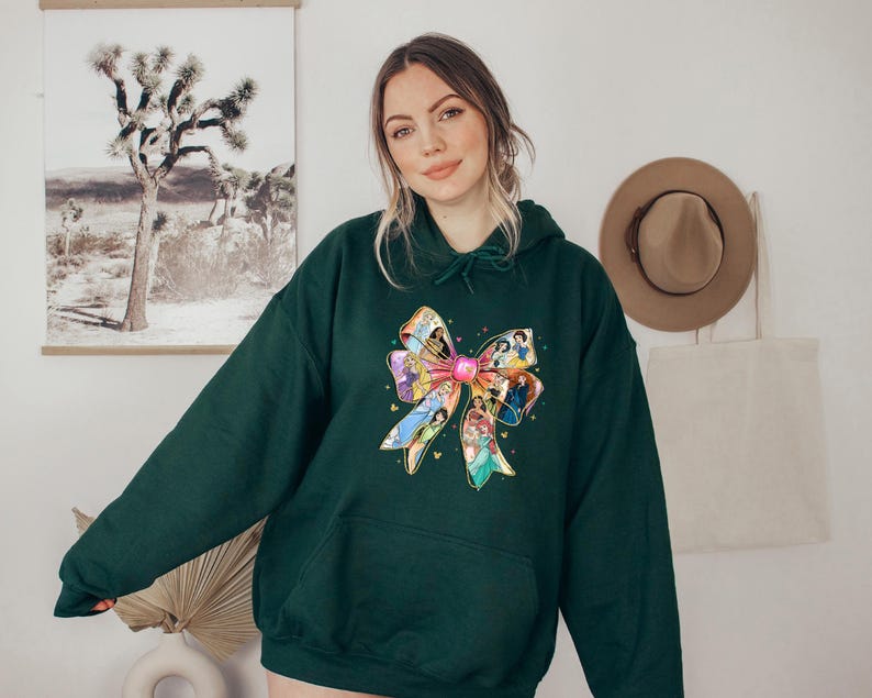 May include: A dark green hoodie featuring a large, colorful bow design. The bow is adorned with images of animated characters. The hoodie has a drawstring hood and a front pocket, offering a casual and comfortable style.