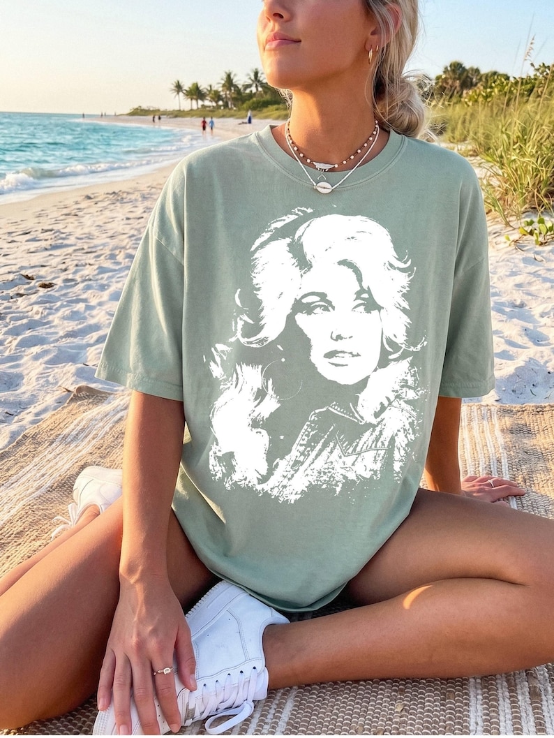 May include: A sage green short-sleeved t-shirt featuring a large, white, high-contrast graphic print of a woman with voluminous hair on the front. The shirt has a crew neckline and a relaxed, loose-fitting silhouette, constructed from cotton fabric.