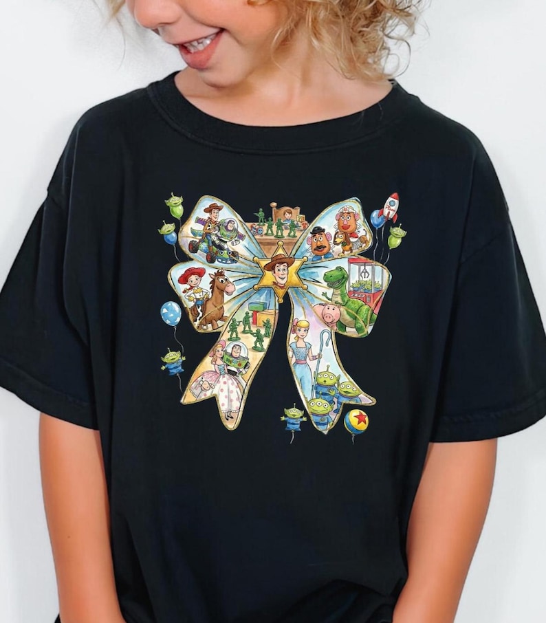 May include: Black t-shirt featuring a colorful graphic of a bow filled with Toy Story characters. The design includes Woody, Buzz Lightyear, and other characters from the animated film. The shirt is made of a soft material.