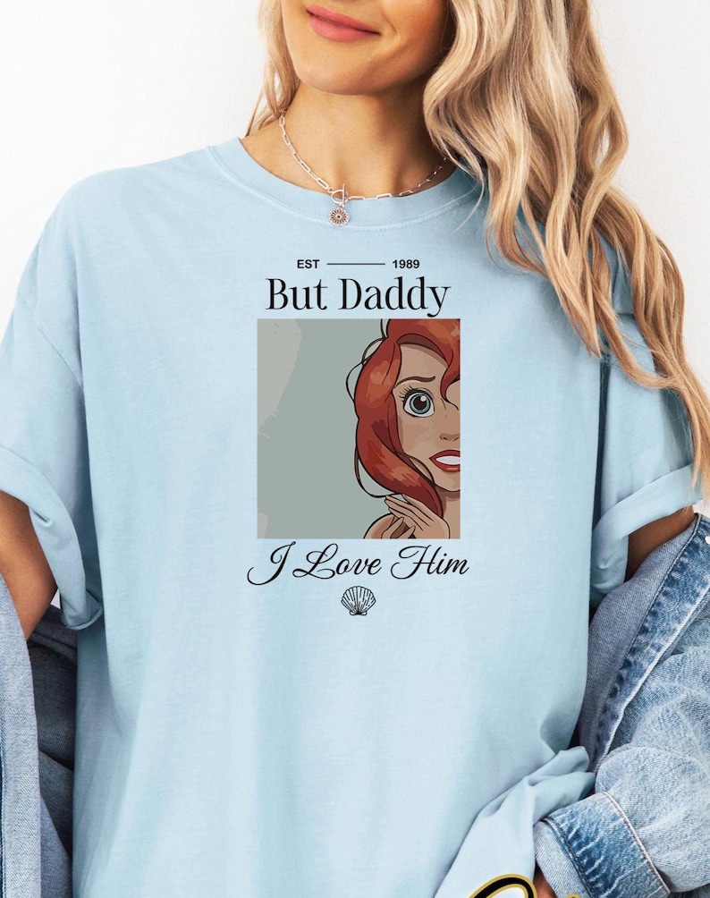 Może przedstawiać: Jasnoniebieska koszulka z napisem "But Daddy I Love Him" i kresk&oacute;wkową ilustracją rudowłosej postaci. Koszulka ma okrągły dekolt i kr&oacute;tkie rękawy. Tekst "EST 1989" znajduje się nad słowami "But Daddy".