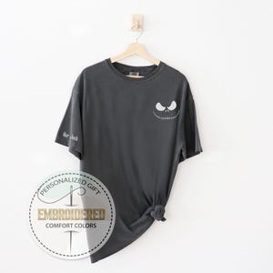 K&ouml;nnte beinhalten: Dunkelgraues T-Shirt mit einem Jack-Skellington-Gesicht auf der Vorderseite und dem Text "Her Jack" auf dem &Auml;rmel. Das T-Shirt h&auml;ngt an einem Holzb&uuml;gel vor einem wei&szlig;en Hintergrund. Das Shirt hat ein rundes Logo mit der Aufschrift "Personalized Gift Embroidered Comfort Colors".