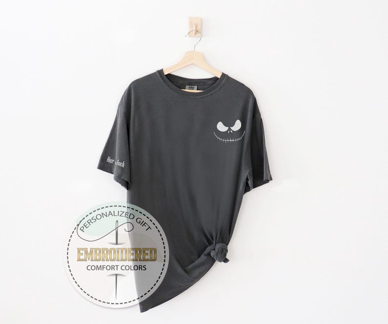 K&ouml;nnte beinhalten: Dunkelgraues T-Shirt mit einem Jack-Skellington-Gesicht auf der Vorderseite und dem Text "Her Jack" auf dem &Auml;rmel. Das T-Shirt h&auml;ngt an einem Holzb&uuml;gel vor einem wei&szlig;en Hintergrund. Das Shirt hat ein rundes Logo mit der Aufschrift "Personalized Gift Embroidered Comfort Colors".