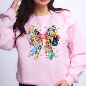 May include: A light pink sweatshirt featuring a large bow design. The bow is filled with images of various princesses. The bow has gold glitter accents and a pink center. The sweatshirt is a solid light pink color.
