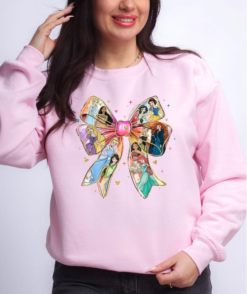 May include: A light pink sweatshirt featuring a large bow design. The bow is filled with images of various princesses. The bow has gold glitter accents and a pink center. The sweatshirt is a solid light pink color.