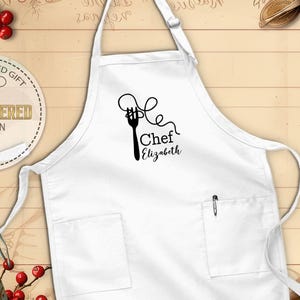 May include: White apron with two pockets and a pen holder. The apron features a black graphic of a fork with spaghetti and the text "Chef Elizabeth". The apron is a personalized gift with an embroidered design.