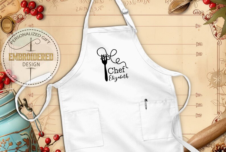 May include: White apron with two pockets and a pen holder. The apron features a black graphic of a fork with spaghetti and the text "Chef Elizabeth". The apron is a personalized gift with an embroidered design.