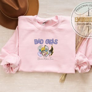 Puede incluir: Una sudadera rosa claro con la frase "BAD GIRLS" en morado y un gr&aacute;fico de tres personajes de dibujos animados. La frase "Have More Fun" est&aacute; debajo. La sudadera forma parte de una colecci&oacute;n de regalos personalizados.