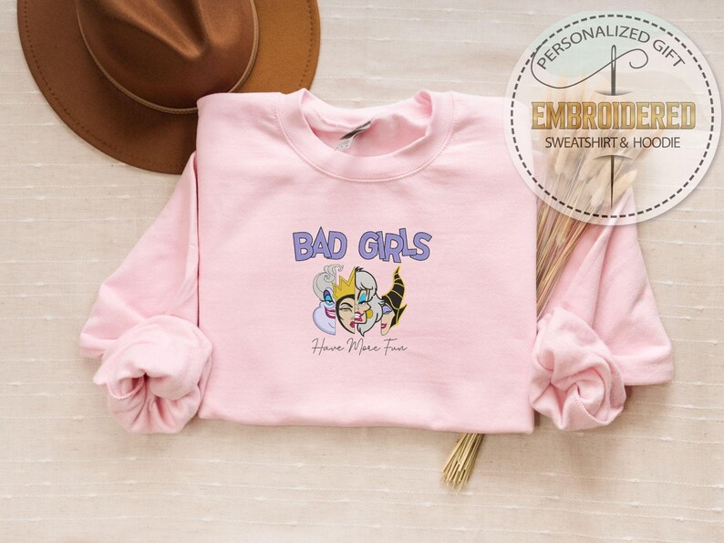 Puede incluir: Una sudadera rosa claro con la frase "BAD GIRLS" en morado y un gr&aacute;fico de tres personajes de dibujos animados. La frase "Have More Fun" est&aacute; debajo. La sudadera forma parte de una colecci&oacute;n de regalos personalizados.