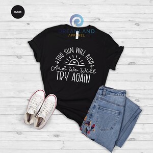 May include: Black t-shirt with a white graphic that says "The sun will rise and we will try again". The graphic includes a stylized sun with a heart in the center.