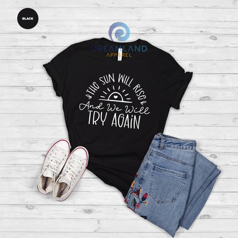 May include: Black t-shirt with a white graphic that says "The sun will rise and we will try again". The graphic includes a stylized sun with a heart in the center.