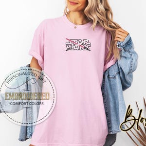 Może przedstawiać: R&oacute;żowy T-shirt z napisem "STAR WARS" czarnymi literami. Za napisem znajdują się dwa skrzyżowane miecze świetlne, jeden r&oacute;żowy, a drugi biały.