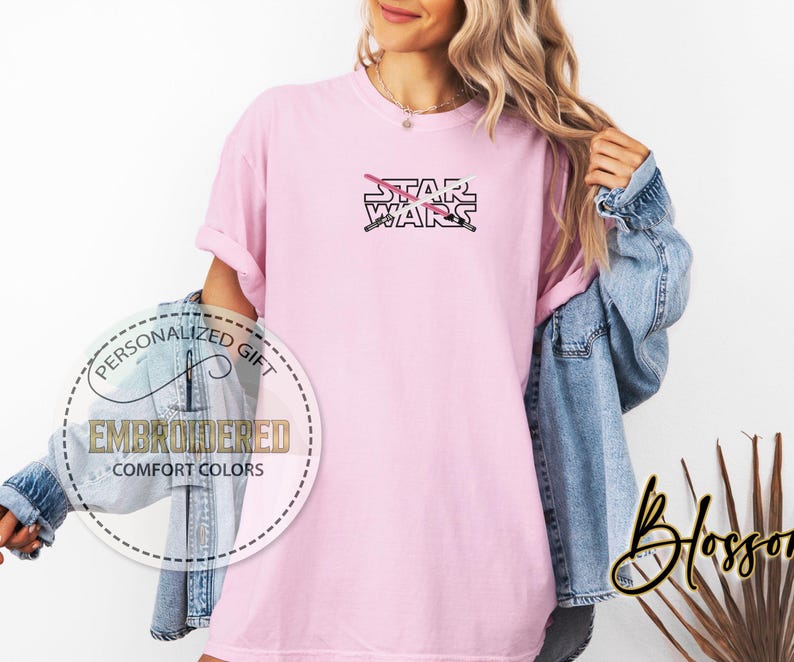 Może przedstawiać: R&oacute;żowy T-shirt z napisem "STAR WARS" czarnymi literami. Za napisem znajdują się dwa skrzyżowane miecze świetlne, jeden r&oacute;żowy, a drugi biały.