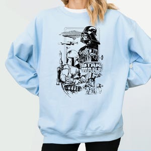 May include: Light blue sweatshirt featuring a black and white Star Wars graphic. The graphic includes Darth Vader, Boba Fett, and the Star Wars logo. The sweatshirt has long sleeves and a crew neck.