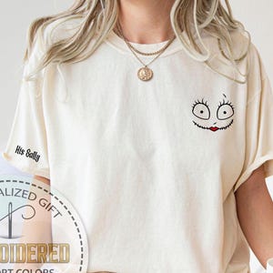 K&ouml;nnte beinhalten: Cremefarbenes T-Shirt mit einem Sally-Gesicht-Motiv auf der Brust und dem Text "His Sally" auf dem &Auml;rmel. Das Sally-Gesicht hat schwarze Stichdetails, gro&szlig;e Augen und einen roten Stichmund. Das Shirt hat eine lockere Passform.