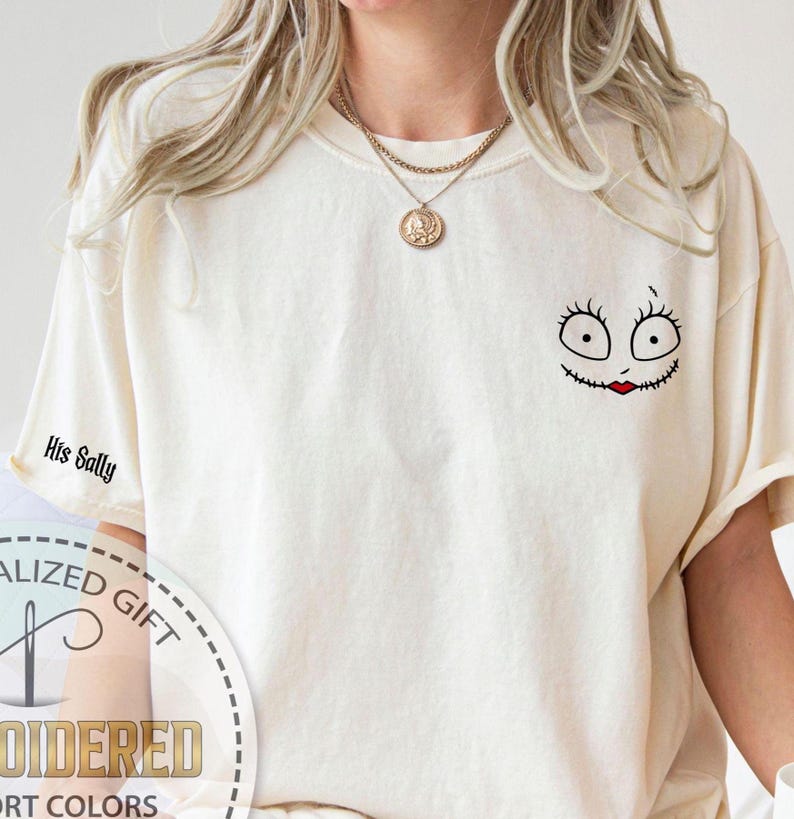 K&ouml;nnte beinhalten: Cremefarbenes T-Shirt mit einem Sally-Gesicht-Motiv auf der Brust und dem Text "His Sally" auf dem &Auml;rmel. Das Sally-Gesicht hat schwarze Stichdetails, gro&szlig;e Augen und einen roten Stichmund. Das Shirt hat eine lockere Passform.