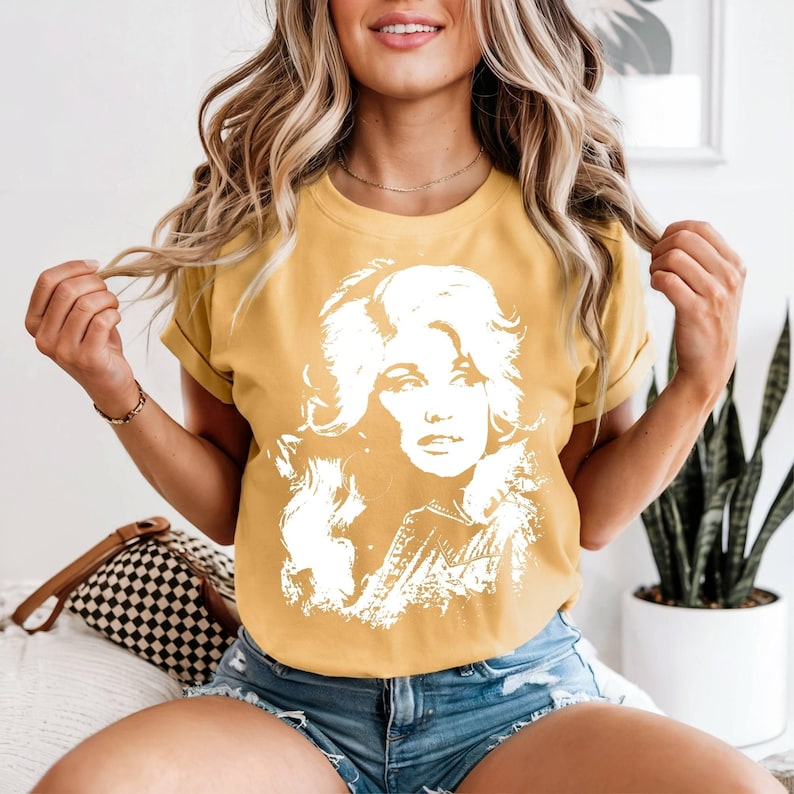 May include: A mustard yellow short-sleeve t-shirt featuring a large, white, high-contrast graphic print of a woman with voluminous hair on the front. The shirt has a crew neckline and rolled sleeves, providing a casual and relaxed fit.