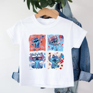 May include: White t-shirt featuring a blue cartoon character in four patriotic designs. The character is wearing a hat, holding a flag, and surrounded by stars and stripes. The shirt is hanging on a wooden hanger.