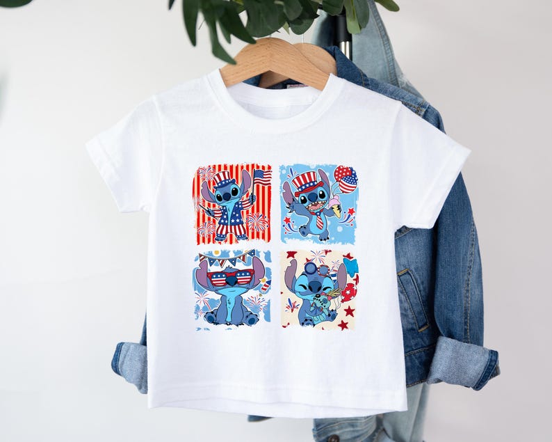 May include: White t-shirt featuring a blue cartoon character in four patriotic designs. The character is wearing a hat, holding a flag, and surrounded by stars and stripes. The shirt is hanging on a wooden hanger.