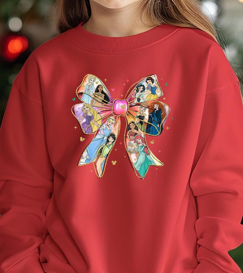 May include: Red sweatshirt featuring a large bow design with various princess illustrations. The bow has a gold outline and a pink center. The princesses are depicted in their iconic outfits, adding a colorful touch to the garment.