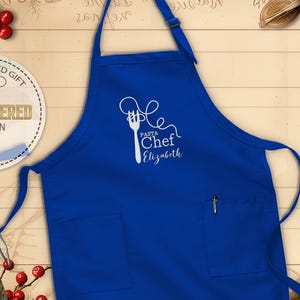 May include: A royal blue apron with a white embroidered design featuring a fork and spaghetti, with the words "Pasta Chef Elizabeth". The apron has two front pockets and an adjustable neck strap. The apron is a personalized gift.