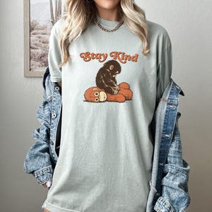 May include: A light green t-shirt with the text "Stay Kind" in orange script. The shirt features a graphic of a brown monkey hugging a red plush toy. The shirt is made of a soft material.