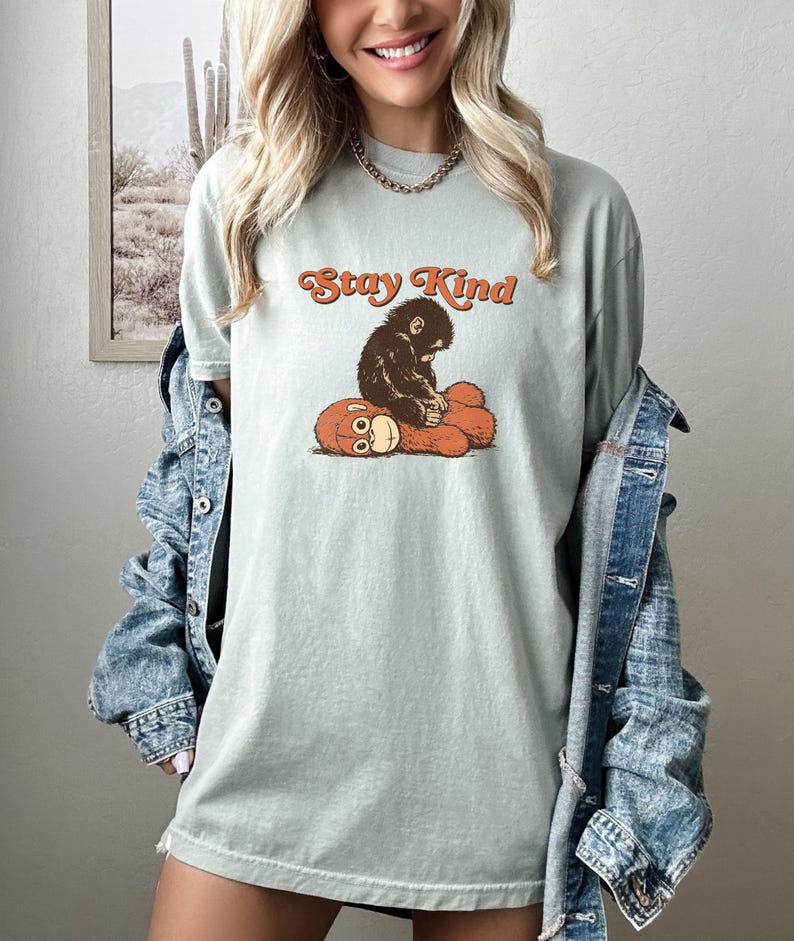 May include: A light green t-shirt with the text "Stay Kind" in orange script. The shirt features a graphic of a brown monkey hugging a red plush toy. The shirt is made of a soft material.