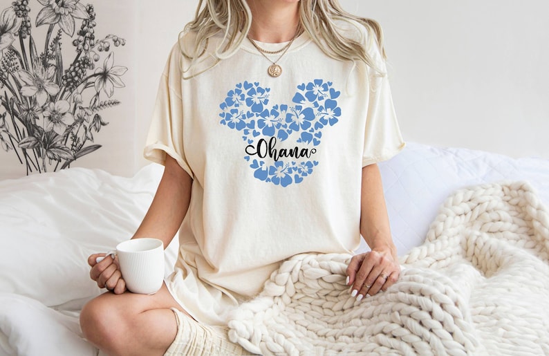Puede incluir: Una camiseta blanca con un dise&ntilde;o de Mickey Mouse floral azul y la palabra "Ohana" en texto negro.