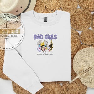 Puede incluir: Sudadera blanca con el texto "BAD GIRLS" en morado y una ilustraci&oacute;n de tres villanos de dibujos animados. Debajo del texto est&aacute; la frase "Have More Fun". La sudadera se muestra sobre un fondo tejido.
