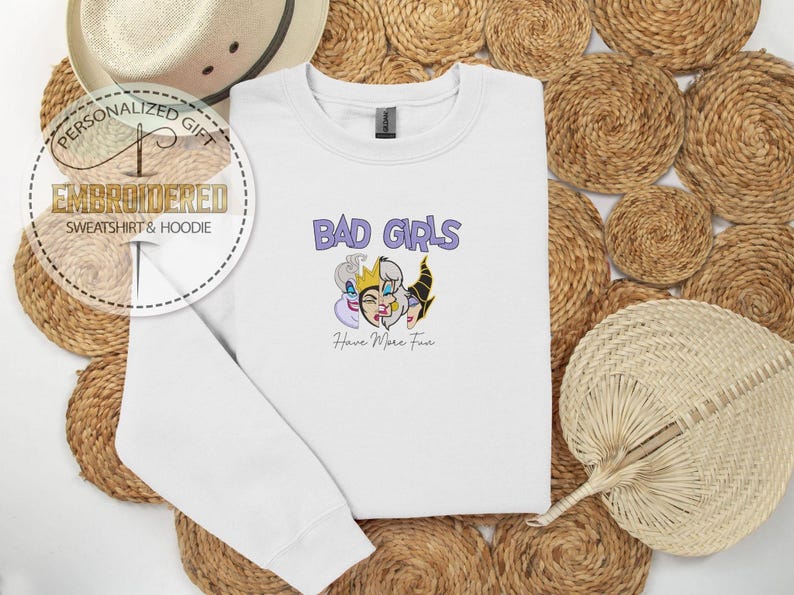 Puede incluir: Sudadera blanca con el texto "BAD GIRLS" en morado y una ilustraci&oacute;n de tres villanos de dibujos animados. Debajo del texto est&aacute; la frase "Have More Fun". La sudadera se muestra sobre un fondo tejido.