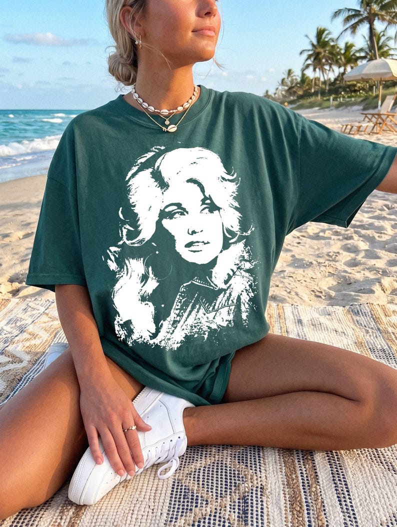 May include: A dark green short-sleeve t-shirt featuring a large, white, high-contrast graphic print of a woman with voluminous hair on the front. The shirt has a crew neckline and a relaxed, loose fit, constructed from a solid-colored cotton fabric.