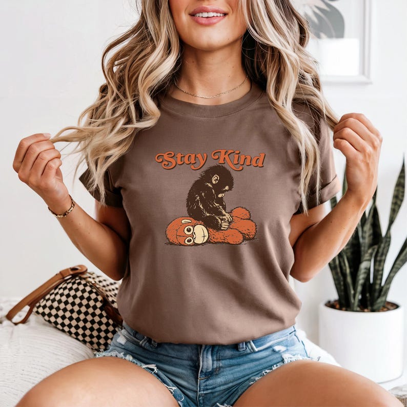 May include: Brown t-shirt with the text "Stay Kind" in orange script. The graphic features a black monkey sitting on a brown teddy bear. The shirt is a casual wear item.
