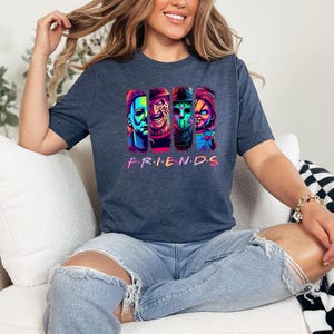 May include: Dark blue t-shirt featuring a colorful graphic of four horror movie characters. The word "FRIENDS" is printed in a rainbow font below the characters. The shirt is made of a soft material.