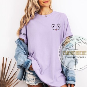 K&ouml;nnte beinhalten: Helllila T-Shirt mit gesticktem Sally-Gesicht-Design auf der Vorderseite. Der &Auml;rmel zeigt den Text "His Sally". Das Shirt geh&ouml;rt zur Comfort Colors Kollektion.