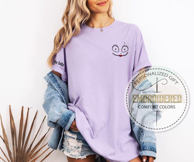 K&ouml;nnte beinhalten: Helllila T-Shirt mit gesticktem Sally-Gesicht-Design auf der Vorderseite. Der &Auml;rmel zeigt den Text "His Sally". Das Shirt geh&ouml;rt zur Comfort Colors Kollektion.