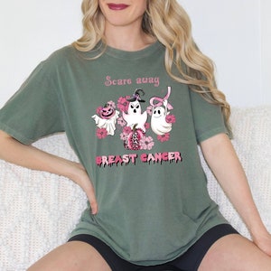 Pode incluir: Camiseta verde com uma fita rosa, fantasmas e ab&oacute;boras. O texto na camiseta diz "Scare away Breast Cancer".