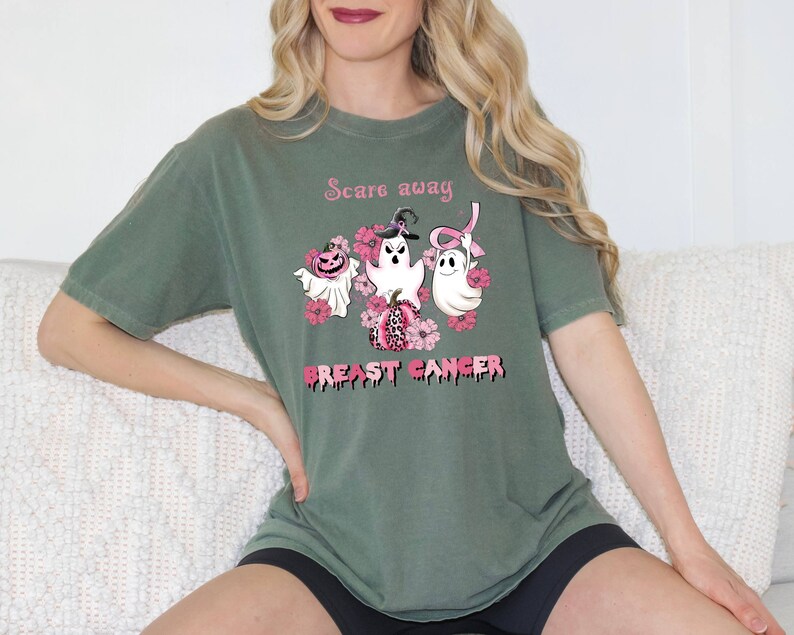 Pode incluir: Camiseta verde com uma fita rosa, fantasmas e ab&oacute;boras. O texto na camiseta diz "Scare away Breast Cancer".
