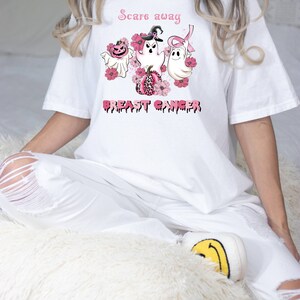 Pode incluir: Camiseta branca com uma fita rosa, fantasmas e ab&oacute;boras. O texto "Scare away breast cancer" est&aacute; impresso na camiseta.