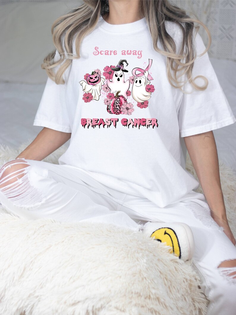 Pode incluir: Camiseta branca com uma fita rosa, fantasmas e ab&oacute;boras. O texto "Scare away breast cancer" est&aacute; impresso na camiseta.