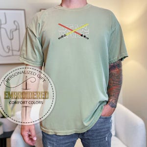 Może przedstawiać: Jasnozielony t-shirt z haftem z napisem "STAR WARS" z przodu. Tekst jest biały z dwoma skrzyżowanymi mieczami świetlnymi w kolorze czerwonym i ż&oacute;łtym.