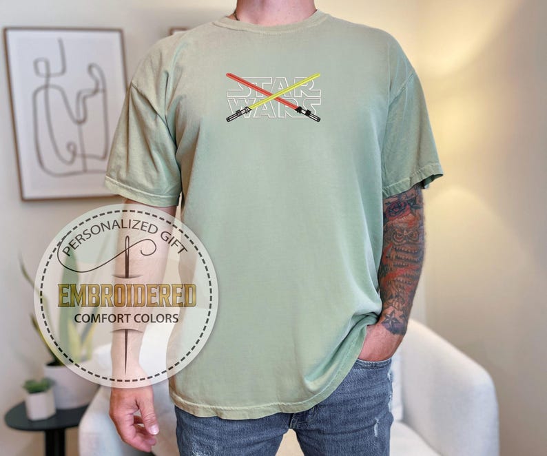 Może przedstawiać: Jasnozielony t-shirt z haftem z napisem "STAR WARS" z przodu. Tekst jest biały z dwoma skrzyżowanymi mieczami świetlnymi w kolorze czerwonym i ż&oacute;łtym.