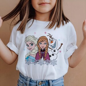May include: White t-shirt featuring Elsa, Anna, and Olaf from the movie Frozen. The graphic includes the characters' faces, snowflakes, and stars. The shirt is worn by a person.
